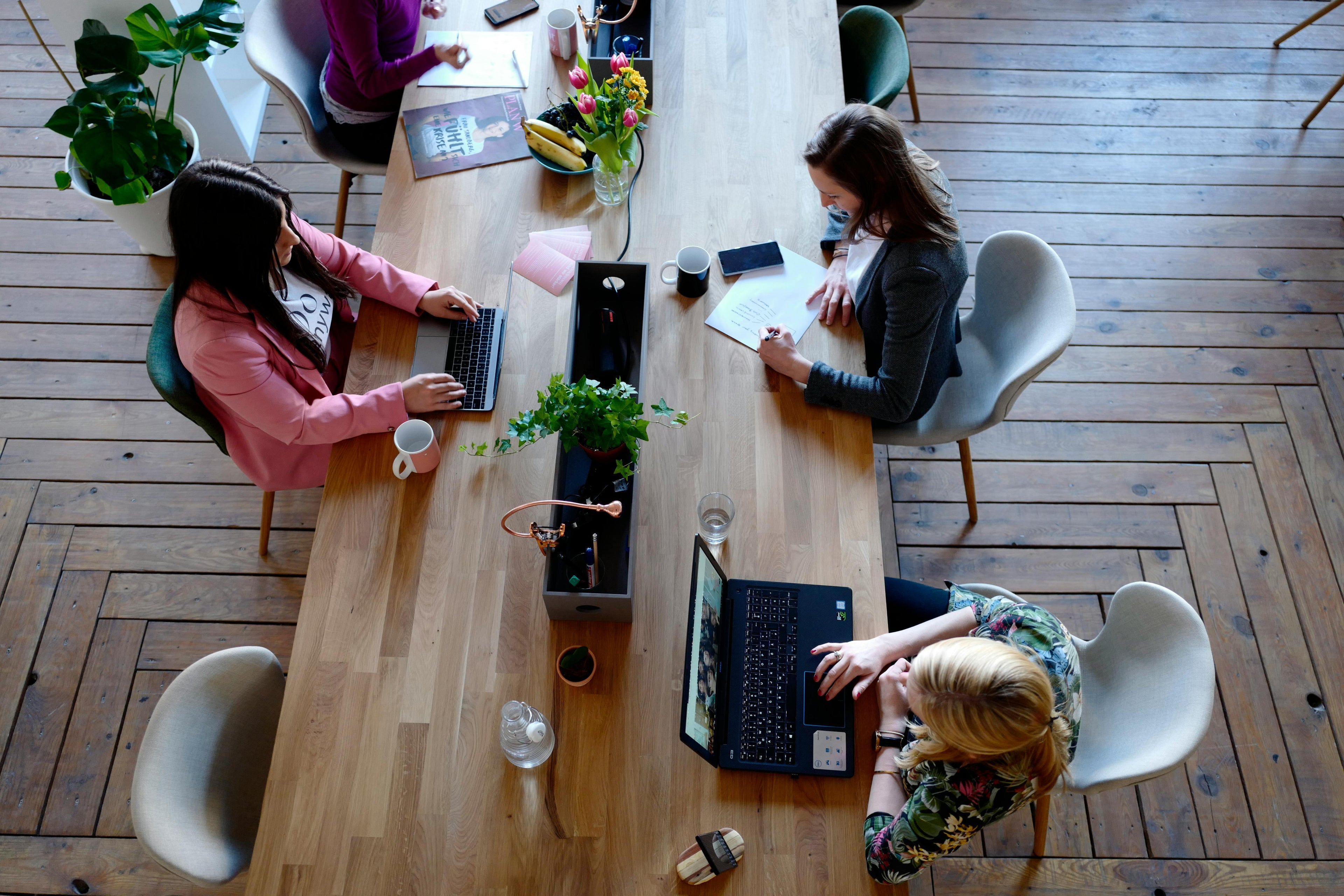 Overhead view of four women collaborating around a long wooden table in a bright office with a herringbone wood floor.